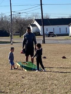 A man and two kids stand around a cornhole set outdoors.