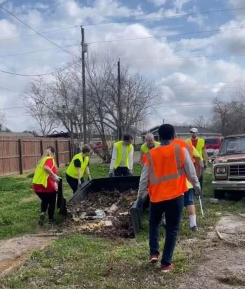 Seven volunteers wearing safety vests hauling a large black container of debris to the street curb